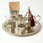 Complete hand-hammered silver plated tea set arranged neatly beside a kraft paper pouch of loose leaf green tea on a clean white surface, showcasing the traditional brewing presentation.