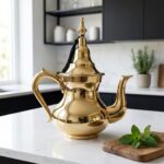 An elegant handcrafted brass teapot displayed on a bright white quartz countertop in a modern Scandinavian-style kitchen with minimalist black cabinetry and fresh greenery.