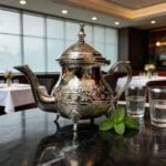 moroccan silver engraved teapot footed on dark reflective surface with a traditional glass of mint tea