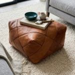 Close-up of a brown square leather floor pouf acting as a coffee table on a textured cream rug in a cozy, contemporary lounge area.