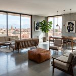 A tan square leather floor pouf centered in a modern, sunlit living room with wooden furniture, polished concrete floors, and large windows overlooking a Moroccan city skyline.