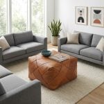 A brown square leather floor pouf styled as a center coffee table on a textured cream rug, surrounded by grey sofas in a bright, contemporary living room.