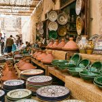 A colorful market stall display filled with various types of Moroccan pottery, including patterned Safi plates, green Tamegroute bowls, and unglazed tagines, under sunlight in a souk.