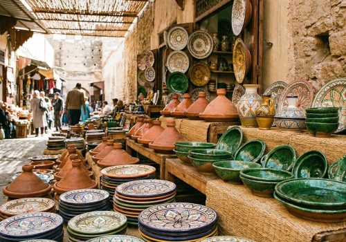 A colorful market stall display filled with various types of Moroccan pottery, including patterned Safi plates, green Tamegroute bowls, and unglazed tagines, under sunlight in a souk.