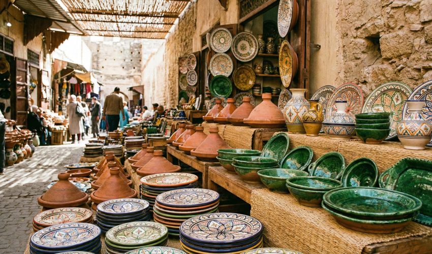 A colorful market stall display filled with various types of Moroccan pottery, including patterned Safi plates, green Tamegroute bowls, and unglazed tagines, under sunlight in a souk.
