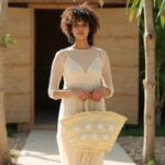 Woman in cream maxi dress holding star straw bag outdoors by pool, handwoven beige tote with white star cotton appliqué