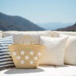 Star straw bag on white outdoor sofa with blue striped cushions, beige handwoven palm leaf tote with white star appliqué