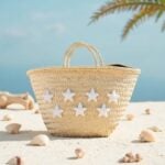 Star straw bag on beach sand surrounded by seashells and driftwood, beige handwoven tote with white star cotton appliqué