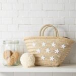 Star straw bag on white shelf beside glass jar with sponges, beige handwoven palm leaf tote with white star appliqué