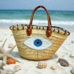Handwoven straw evil eye beach bag with blue nazar embroidery on sandy beach beside seashells