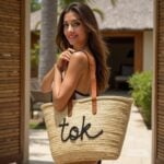 Personalized straw bag with black cursive embroidery worn by woman in tropical setting, handwoven palm leaf