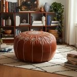 Light brown leather floor ottoman in a cozy room with a geometric rug, bookshelves, and trailing house plants.