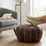 A person resting their feet, wearing cozy beige knit socks, on a dark brown leather floor ottoman next to a gray textured armchair.