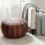 Chocolate brown leather floor ottoman positioned near a window next to a white armchair with a grey knit throw.