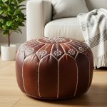 Dark brown leather floor ottoman with white embroidery sitting on a light wood floor next to a white sofa.