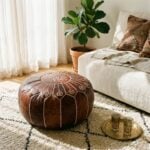 A dark brown leather floor ottoman resting on a plush white patterned rug in a cozy, sunlit living room near a sofa and a brass tray.