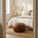 A dark brown leather floor ottoman sitting at the foot of a neatly made bed in a bright, neutral-toned bedroom.