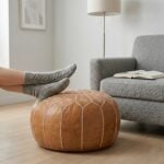 A person relaxing with their feet in gray knit socks propped up on a tan leather floor ottoman beside a modern gray armchair.