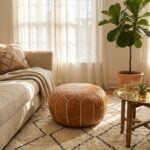 A tan leather floor ottoman styled in a warm, sunny living room on a geometric rug, sitting next to a beige sofa, a fiddle leaf fig, and a brass side table.