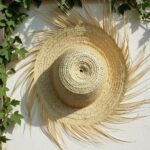 Overhead view of artisanal straw accessories wide brim hat with dramatic palm fringe against white wall and greenery