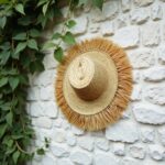 Handwoven straw garden hat with fringe on white stone wall, artisan spiral weave, boho accessories