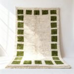 Handwoven Moroccan rug with emerald green square border pattern on cream wool, displayed draped showing plush texture