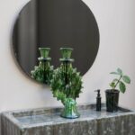 Green ceramic candlestick with drip glaze finish on concrete sink beside round mirror and potted plant