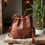 Brown leather backpack with hand-tooled embossed patterns and scalloped mid-band on wooden bench beside green plant