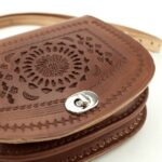 Close-up of hand-tooled sun-ray mandala embossing on cognac brown leather crossbody bag flap with decorative stitching