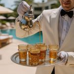 Elegant waiter in a white tuxedo pouring tea from a silver pot into gold Moroccan tea glasses on a round brass tray beside a turquoise pool.