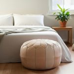 Handcrafted leather floor ottoman at foot of bed in neutral bedroom with white linens and natural light from window