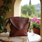 Handmade brown leather crossbody purse with envelope closure on marble table by garden window
