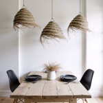 Three woven straw chandeliers above rustic wooden dining table with black chairs