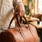 Close-up of hand-stitched leather travel bag handles and brass fittings, held by a person in a linen shirt in a warm café setting