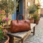 Vintage-style leather travel bag displayed on a rustic wooden bench in a charming Mediterranean courtyard with olive trees and bougainvillea