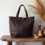 Chocolate brown leather tote bag handcrafted design on rustic wooden table with dried wheat