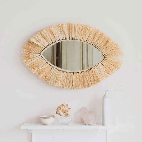 Handwoven straw eye mirror in natural raffia hanging on white wall above minimalist shelf with boho bedroom decor