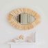 Handwoven straw eye mirror in natural raffia hanging on white wall above minimalist shelf with boho bedroom decor