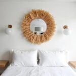 Natural raffia hanging mirror with linen rope above white bed creating serene bohemian coastal bedroom wall decor