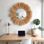 Round suspended straw mirror with rope hanging system above modern workspace desk with monstera leaf in bohemian office
