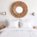 Round handwoven straw mirror centered above white bed with neutral bedding in serene bohemian bedroom design