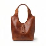 Studio shot of a minimalist brown leather shoulder bag isolated on a white background, featuring a curved hobo-style silhouette and a central vertical strap with a brass buckle.