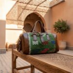Green kilim leather duffle bag with colorful handwoven wool patterns displayed on carved wooden bench in Moroccan setting