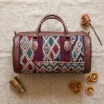 Top-down flat lay of kilim leather duffle bag on linen with brass Moroccan tea glass, dried rose, and orange slices