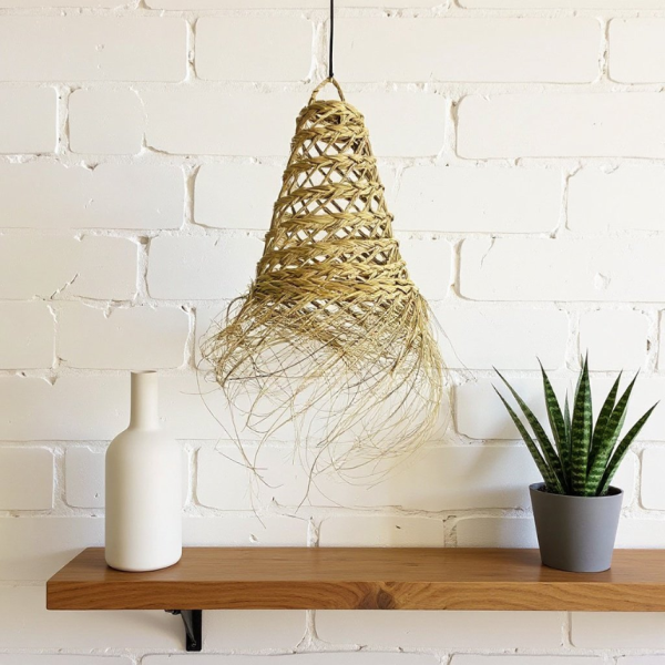 Handmade Moroccan straw light shade with frayed edge hanging above wooden shelf against white brick wall, vase and snake plant decor