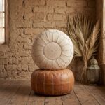 Vertical stack of two Moroccan ottomans, featuring the undyed genuine leather pouf on top of a classic tan leather version against a textured mud wall.