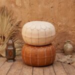 Vertical stack of two Moroccan ottomans, featuring the undyed genuine leather pouf on top of a classic tan leather version against a textured mud wall.
