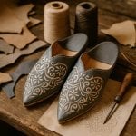 Handcrafted engraved leather babouche slippers in gray with gold arabesque patterns beside crafting tools and thread spools