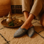 Relaxed scene with blue Moroccan babouche slipper on cushioned rug beside brass tray, silver teapot, and steaming mint tea glasses.
