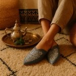 Person wearing sage green engraved leather babouche with floral arabesque pattern beside brass Moroccan tea set on rug